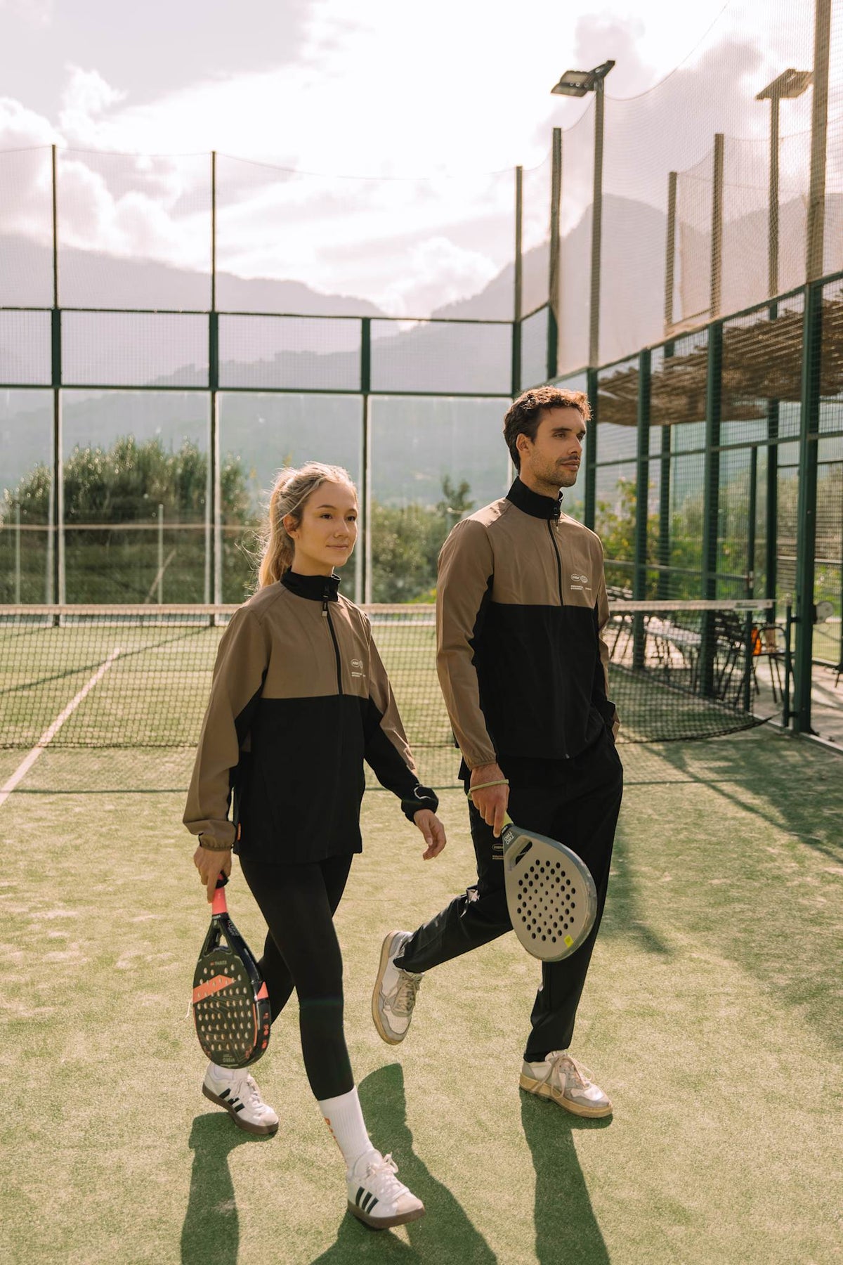 Model wearing Cuera Active Track Jacket in Black / Walnut, lifestyle action shot on the padel court